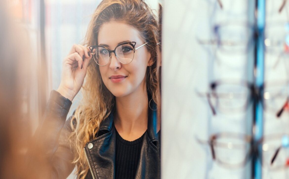 Smiling young woman trying on glasses on mirror in optician.