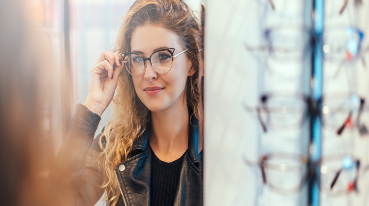 Smiling young woman trying on glasses on mirror in optician.